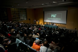 A wide shot image of filled auditorium and five speakers seated at stage