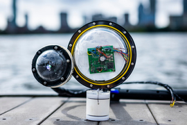 A prototype camera, with 2 bulbous lenses, a circuit board, wires and white cylinder, sits on a dock with the Charles River and Boston skyline in blurred background. 