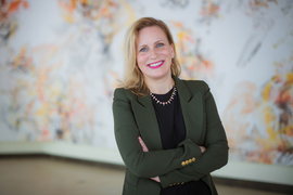 Photo of Angela Koehler smiling with arms crossed. Indoors with white, yellow, and orange mural in background.