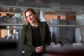 Angela Koehler inside the lab, with blurry foreground of lab shelving, and desk and shelves with equipment in background.  