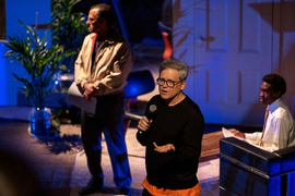 Jay Scheib holds a microphone and addresses the audience while on stage. 2 people are blurry in the background.