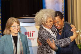 Sally Kornbluth smiles as Angela Davis hugs Melissa Nobles, with podium in background.