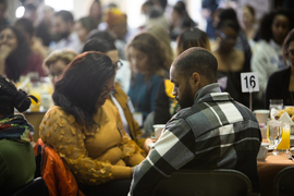 Audience members are seated at tables, with heads bowed in thought and prayer.