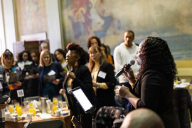 Woman with microphone sings as the audience stands, and some sing along.