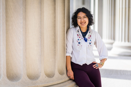 Dogan leans against a beige column while on MIT campus, and wears a Brass Rat ring. More columns are in the background.