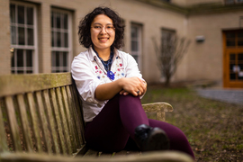 Dogan relaxes on a wooden bench on campus, with hands on knee. A campus building and gardens are seen in the background.