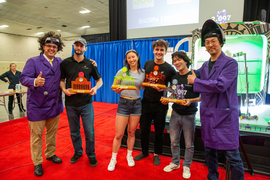 The four winning students and two faculty instructors stand together, smiling. Amos Winter and Sangbae Kim wear purple lab coats. The students each hold laser-cut awards in the shape of the Killian columns and Great Dome.