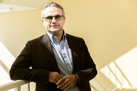 Yilmaz leans on a banister in a stairwell. The background is beige and sunlight creates diagonal shadows on the wall.