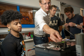 Surrounded by 3 students, Kolle, center, reaches for an object. They are inside a lab.