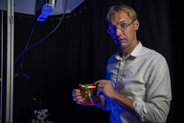 Kolle stands against a black backdrop, lit by a blue light. He holds a stretchy material with Tim the Beaver on it.