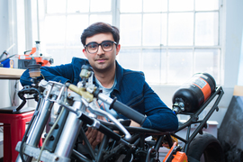 Adi Mehrotra smiles while resting his arms on a handmade motorcycle. Mehrotra is inside a shop and windows in background.