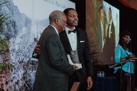 Clarence Williams stands on a stage while holding a plaque and smiling, as Karl Reid has his arm around Williams’ back. A greyscale photo of the 1963 March on Washington is in the background.