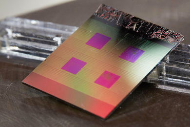 Photo of an integrated circut, a square pane with four smaller magenta squares within, on a brown desktop