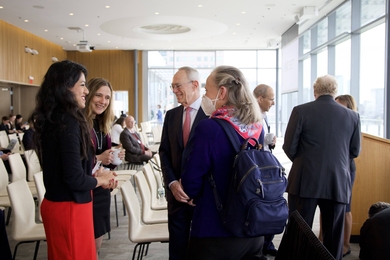 Photo of four standing people conversing in a lecture hall with a wall of windows on the right.