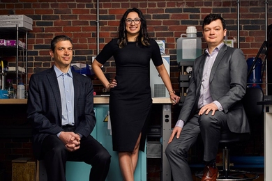 Three people pose in front of a lab bench and a brick wall. Two are seated and one is standing.