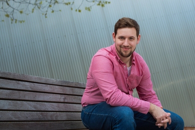 Alexander O'Brien sits leaning on his knee on a wooden bench, with a corrugated metal wall in the background