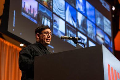 Refik Anadol speaks at the podium, with slide projection in background.