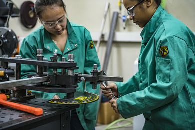 Two young women in teal safety jumpsuits work together in a machine shop